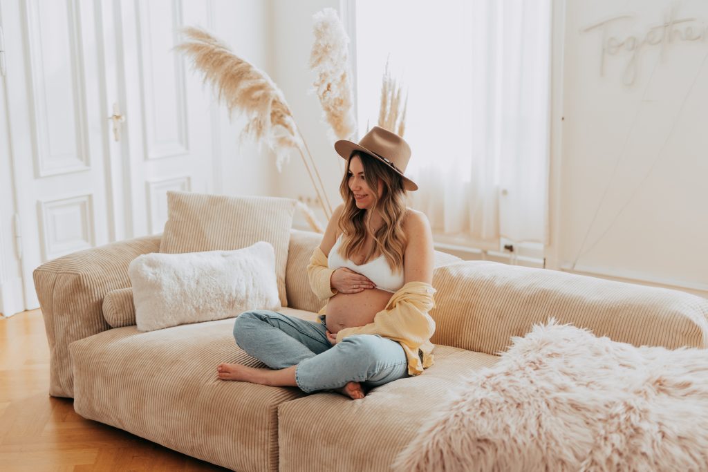 Schwangere Frau sitzt auf einem Sofa und streichelt ihren Bauch.